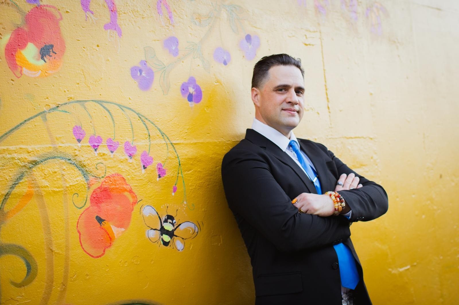 Steve Pinney Jr in a dark suit and blue tie against a yellow painted wall